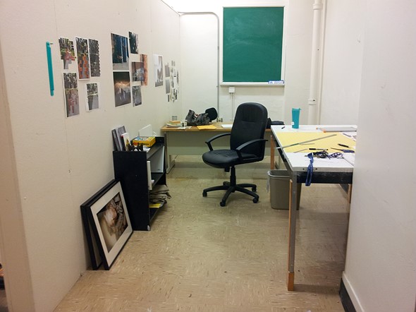 A small studio workspace has photos pinned to the walls  a black office chair  drafting tables and art supplies scattered around the room