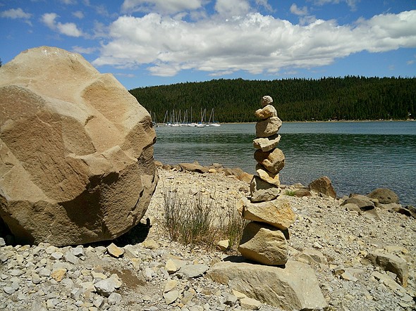Pile de roches équilibrées sur un lac rocheux près d'un gros rocher avec des collines boisées d'eau calme et des voiliers en arrière-plan