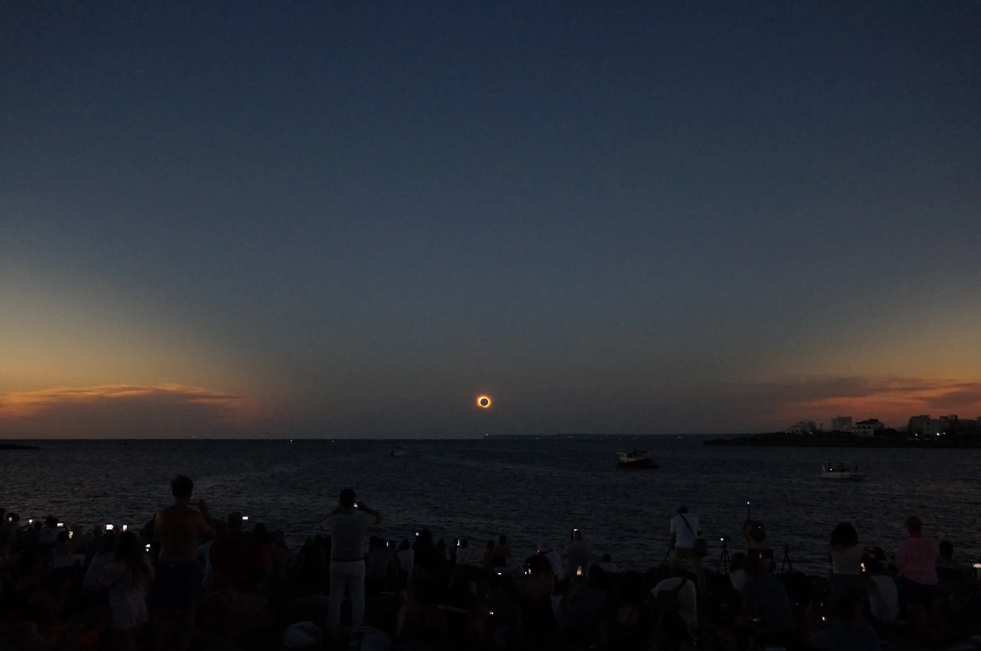 A crowd of people stands on a beach, all holding their phones up and dotting the landscape with tiny pinpricks of light. In the sky, you can see a full eclipse.