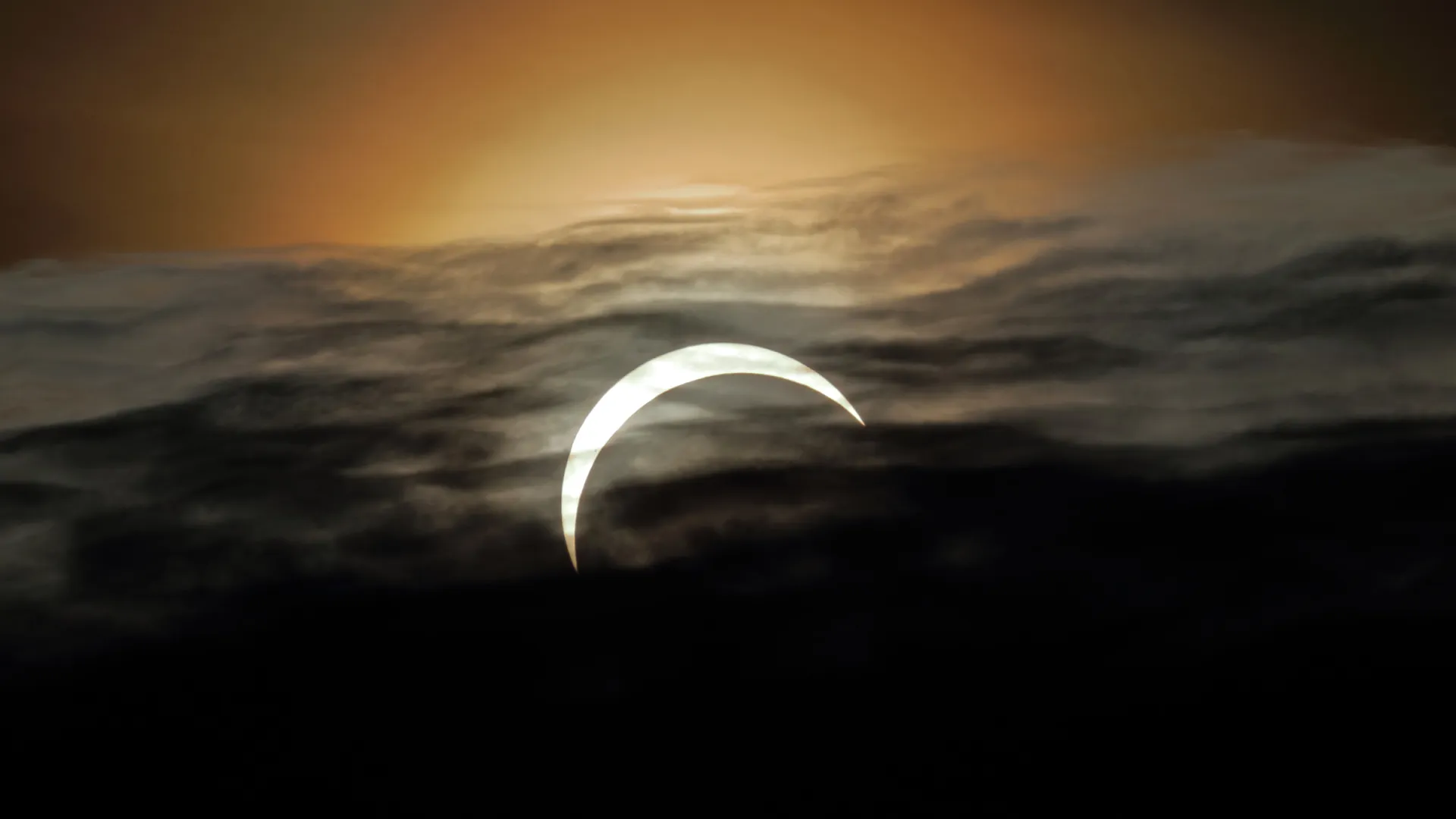 An image of a partial eclipse behind cloud cover. The sky glows a pallid orange above it.