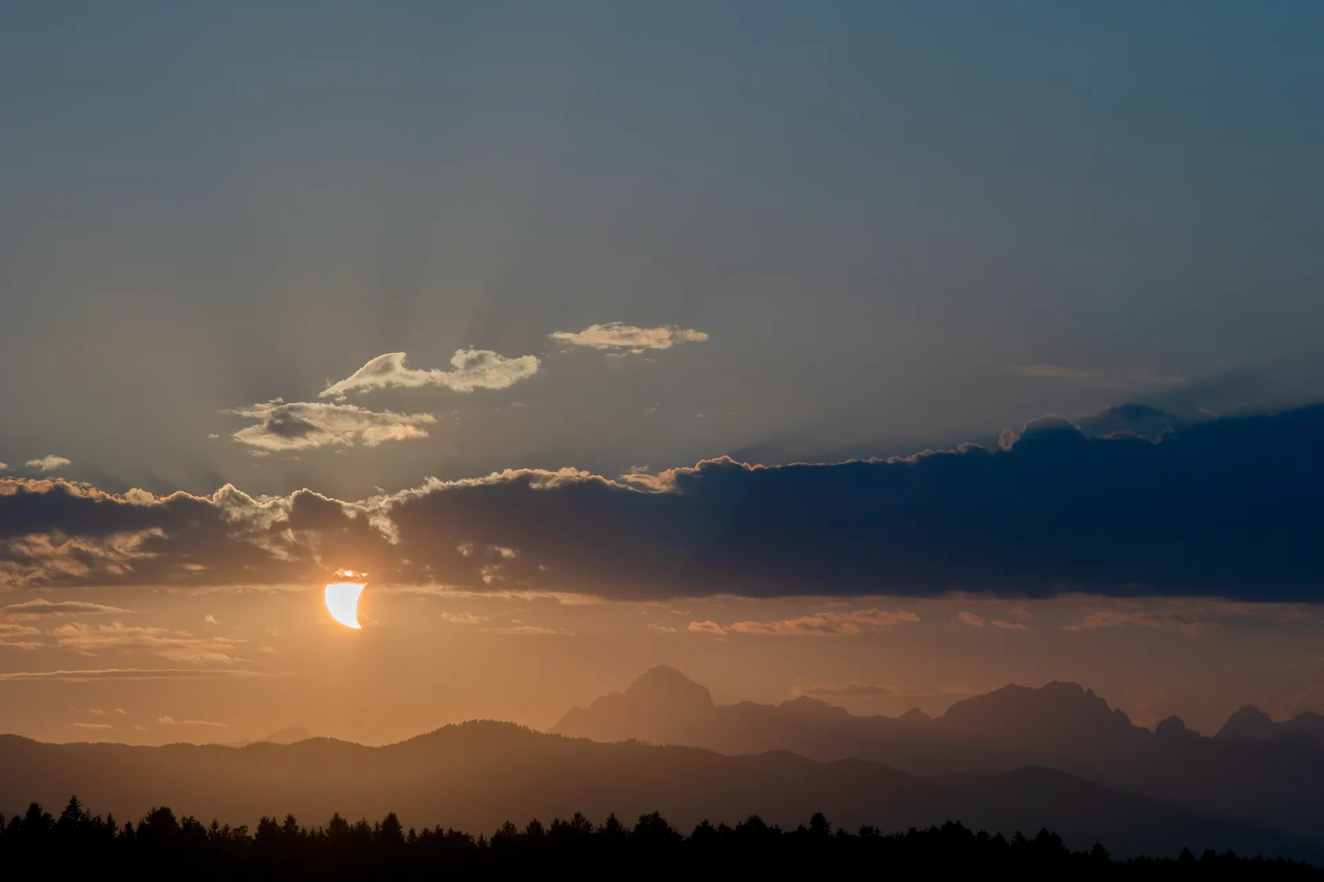 A landscape with trees in the foreground, and layers of mountains in the background. A stripe of clouds splits the sky into orange and blue, and the sun – partially obscured by the moon – peeks out from under them.