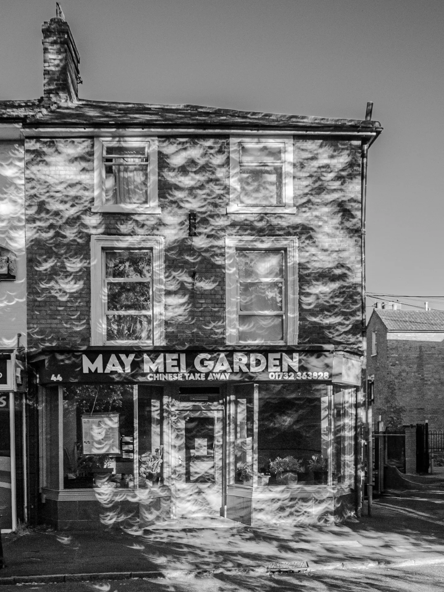 Black and white photo of a restaurant. Its façade looks dimpled, because of the hundreds of projections of a partial eclipse that are falling on it.