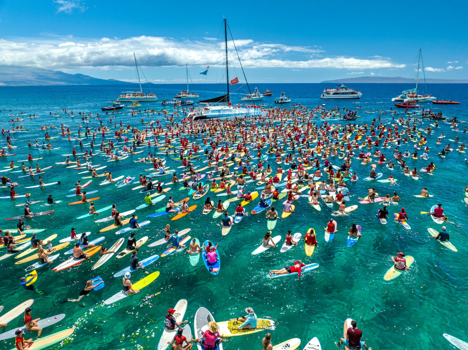 Large group of surfboards clustered in clear blue ocean water near several boats under a sunny sky, forming a vibrant, celebratory scene.