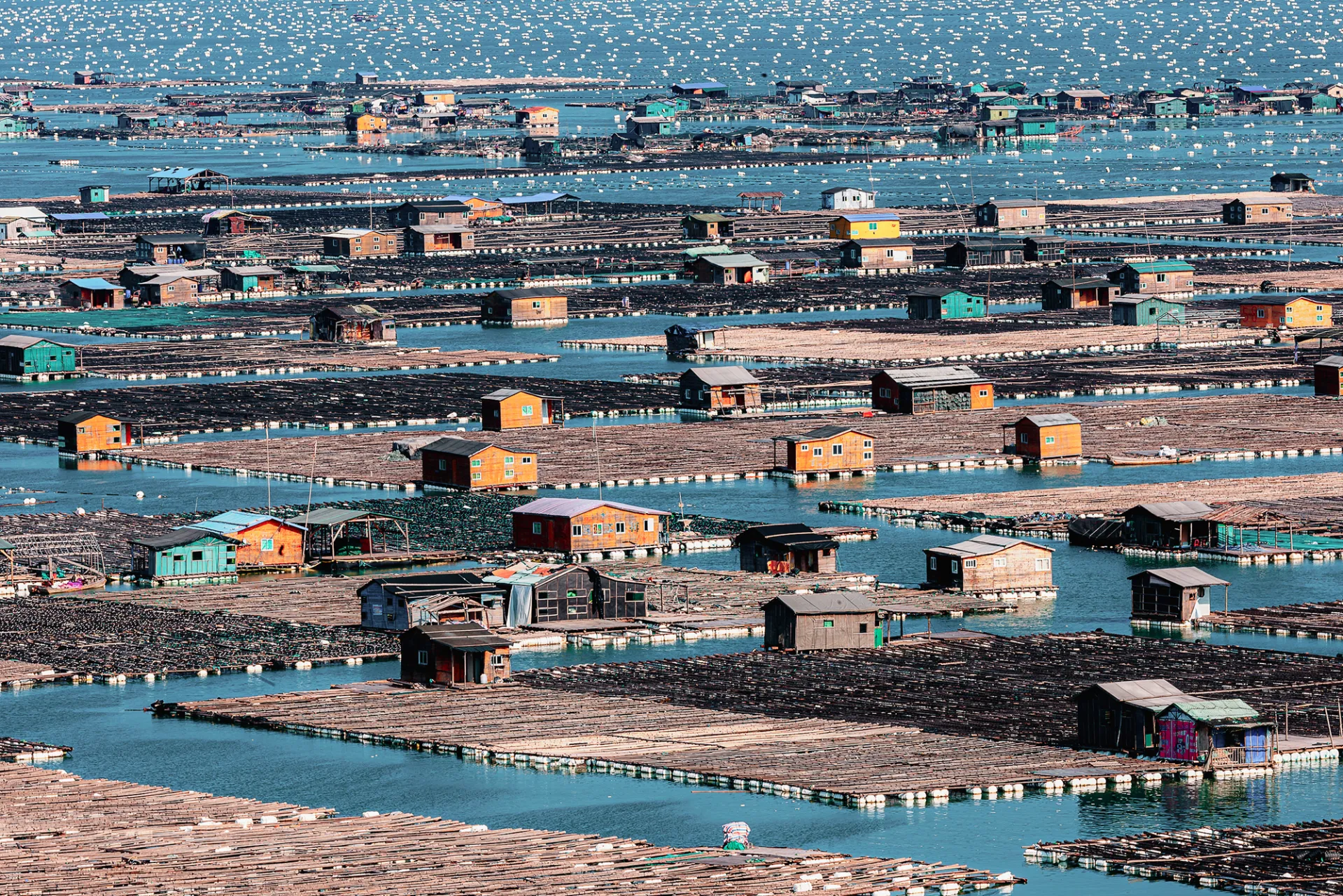 Floating fish farms with rows of colorful huts are positioned on the blue sea, with many small white buoys visible in the background.
