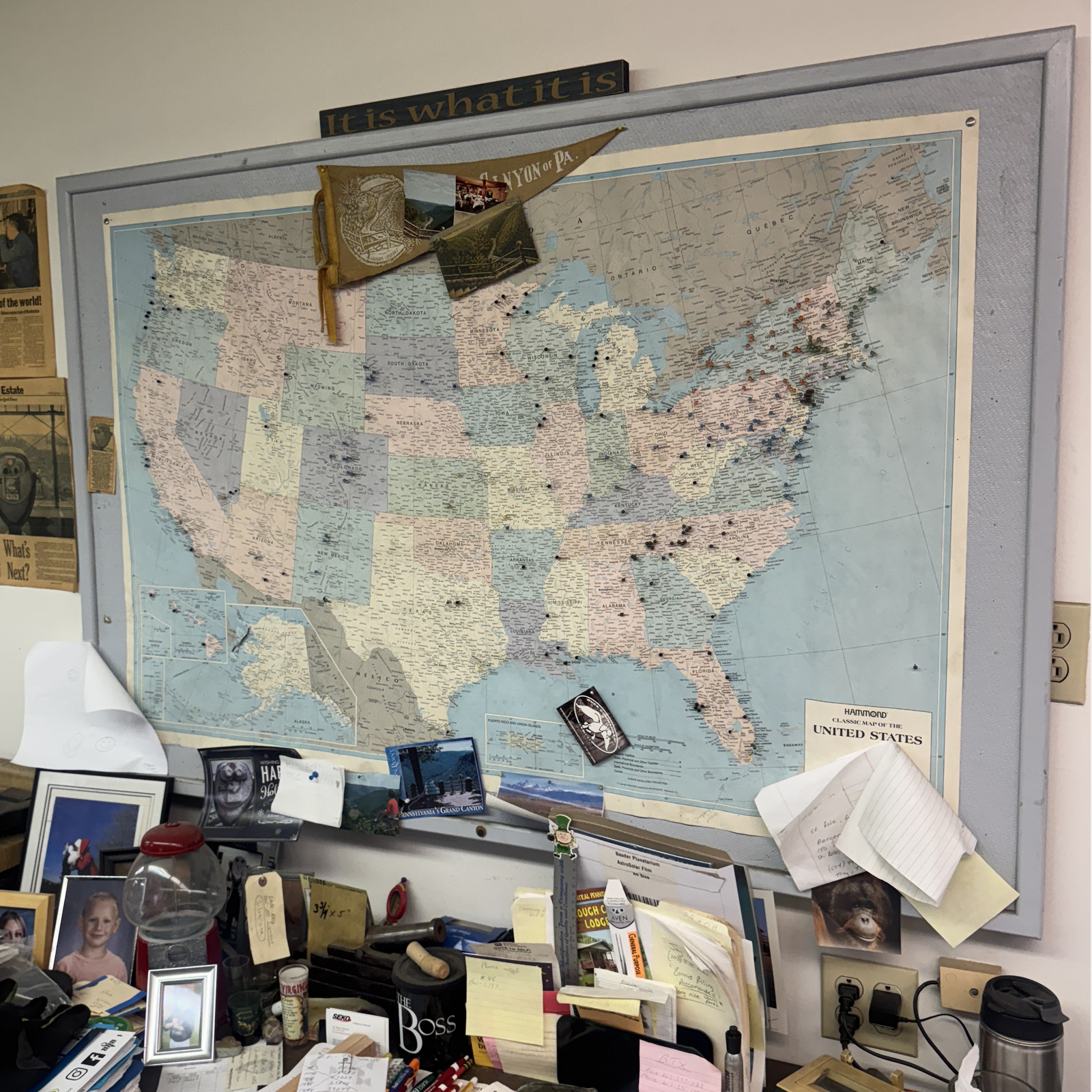 Cluttered desk with scattered papers and photos beneath a large, pinned map of the United States in a molded frame with small pins representing the locations of Tower Optical binocular viewfinders; a sign above the map displays the phrase 