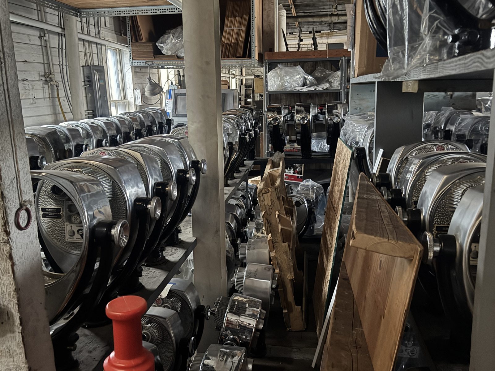 Rows of shiny metal binocular optical viewers are arranged in a dimly lit, cluttered storage room with wooden shelves and assorted industrial equipment.