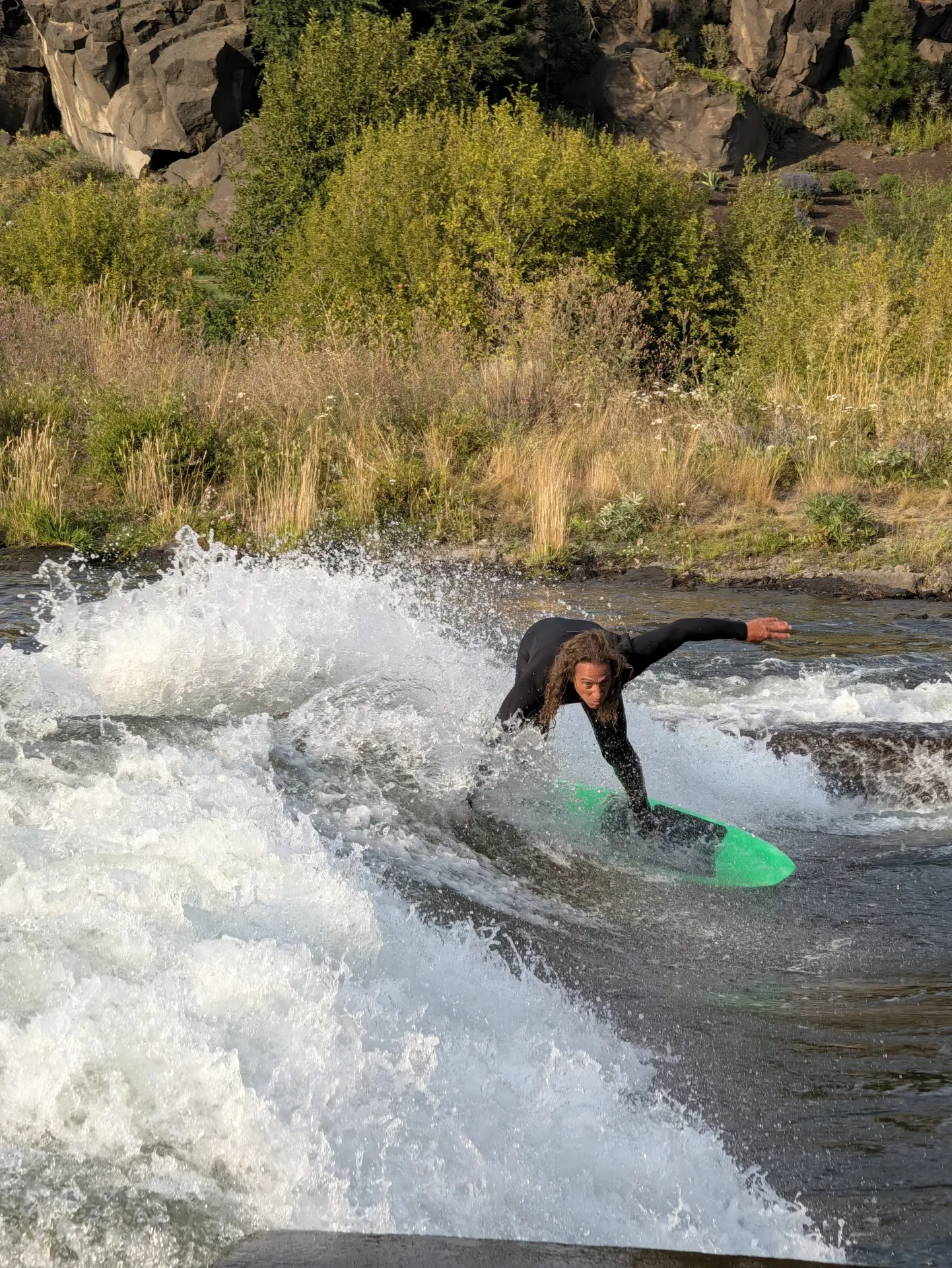 A person in a black wetsuit stands on a bright green surfboard while surfing a river wave, with white water and greenery in the background.
