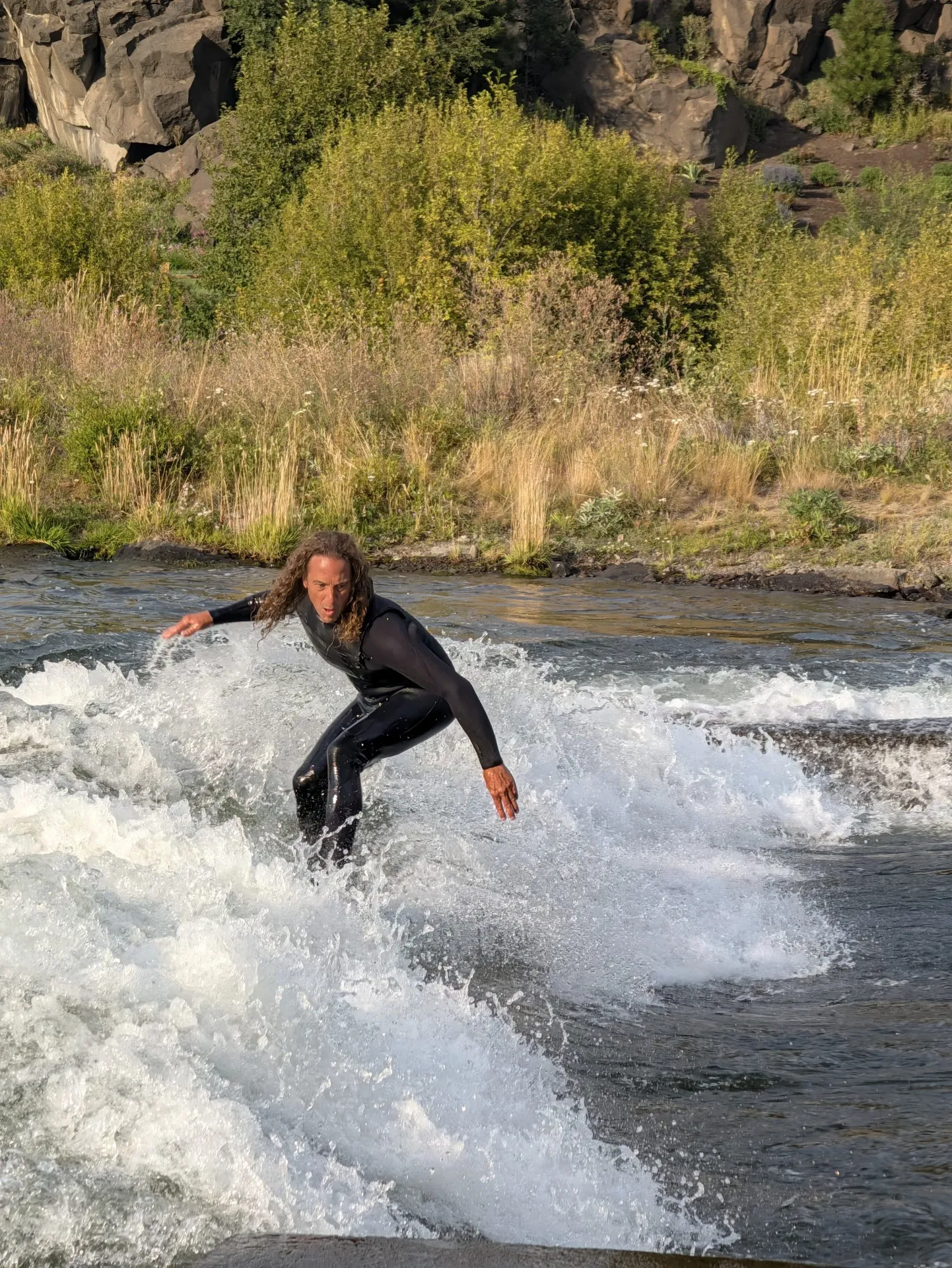 A person in a black wetsuit stands on a bright green surfboard while surfing a river wave, with white water and greenery in the background.