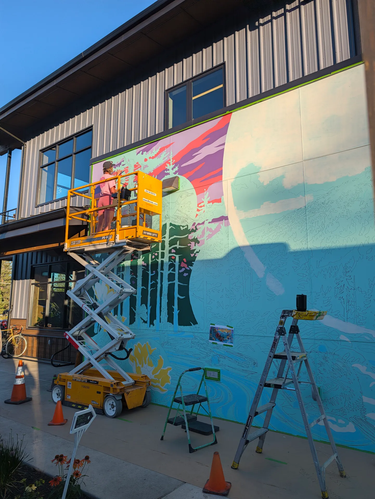 A yellow lift holds an artist painting a vibrant mural of trees and a mountain sunset on an outdoor wall, with a ladder, traffic cones, and bicycle positioned nearby.