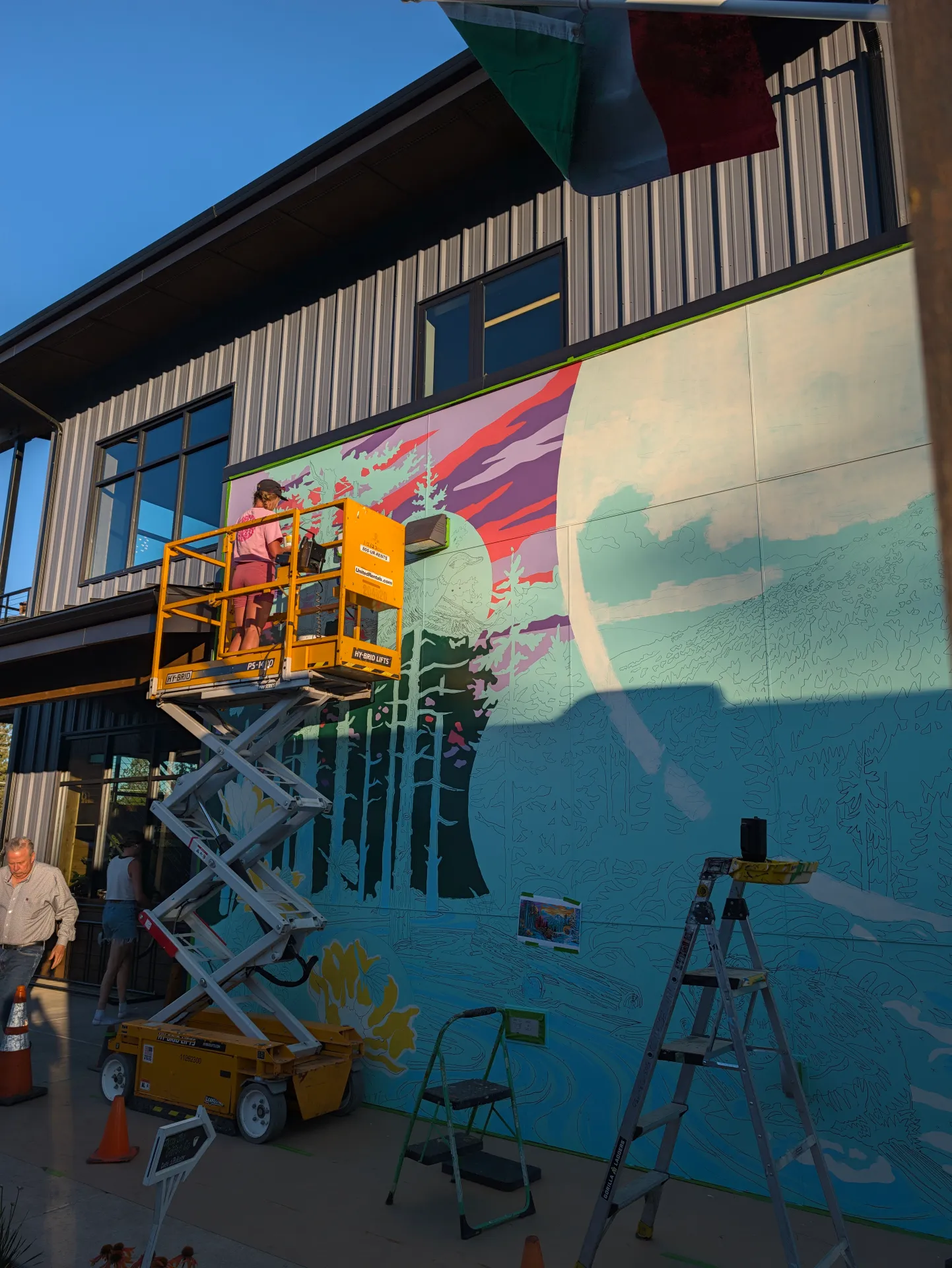 A yellow lift holds an artist painting a vibrant mural of trees and a mountain sunset on an outdoor wall, with a ladder, traffic cones, and bicycle positioned nearby.