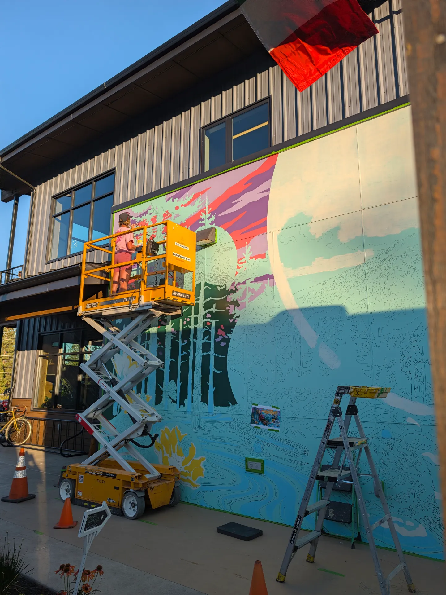 A yellow lift holds an artist painting a vibrant mural of trees and a mountain sunset on an outdoor wall, with a ladder, traffic cones, and bicycle positioned nearby.