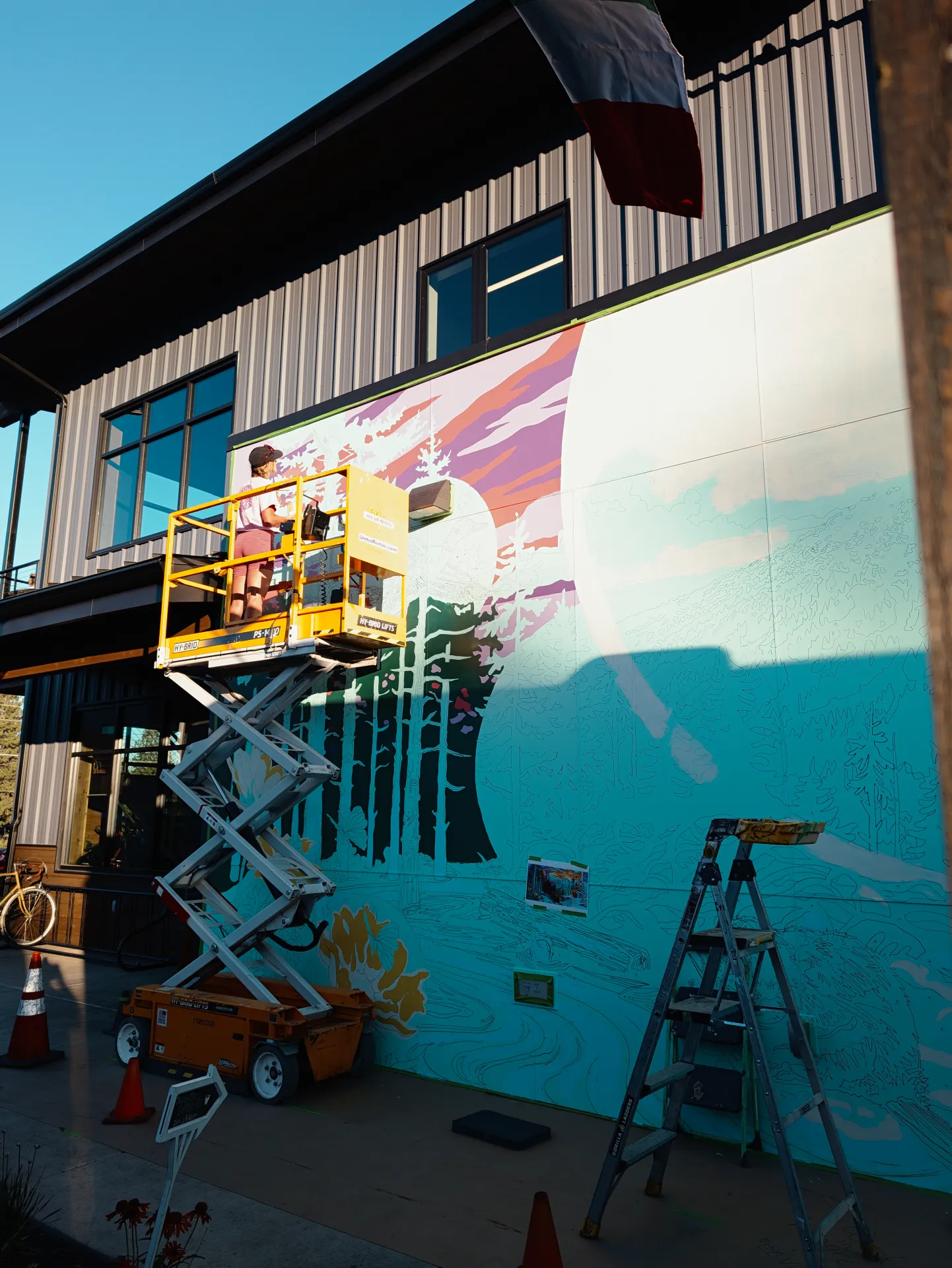 A yellow lift holds an artist painting a vibrant mural of trees and a mountain sunset on an outdoor wall, with a ladder, traffic cones, and bicycle positioned nearby.