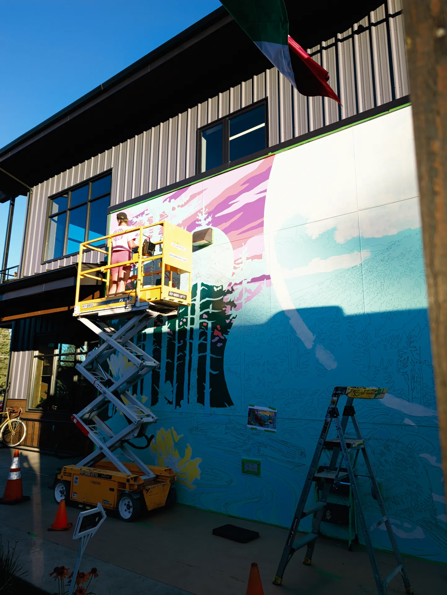 A yellow lift holds an artist painting a vibrant mural of trees and a mountain sunset on an outdoor wall, with a ladder, traffic cones, and bicycle positioned nearby.