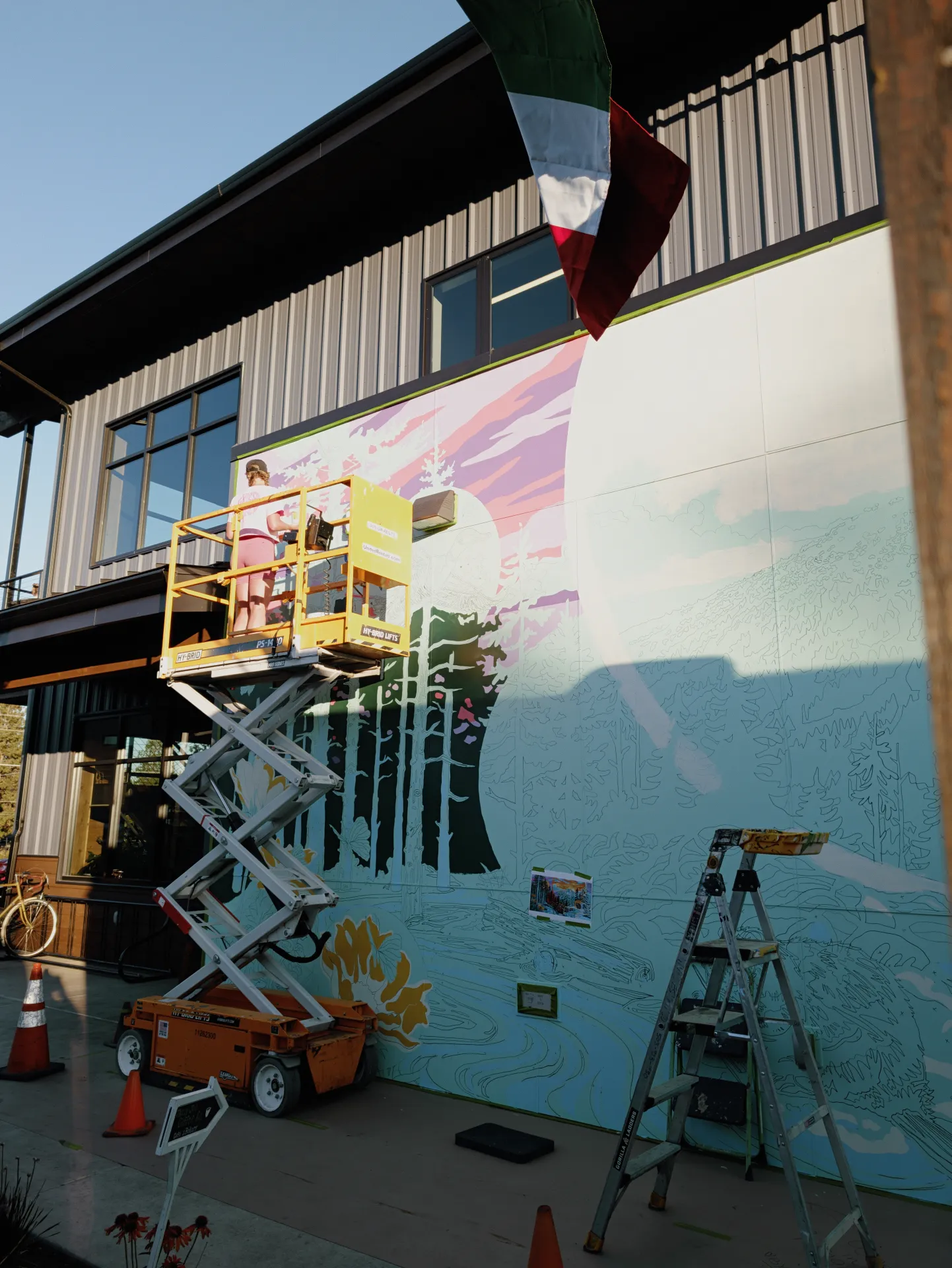 A yellow lift holds an artist painting a vibrant mural of trees and a mountain sunset on an outdoor wall, with a ladder, traffic cones, and bicycle positioned nearby.