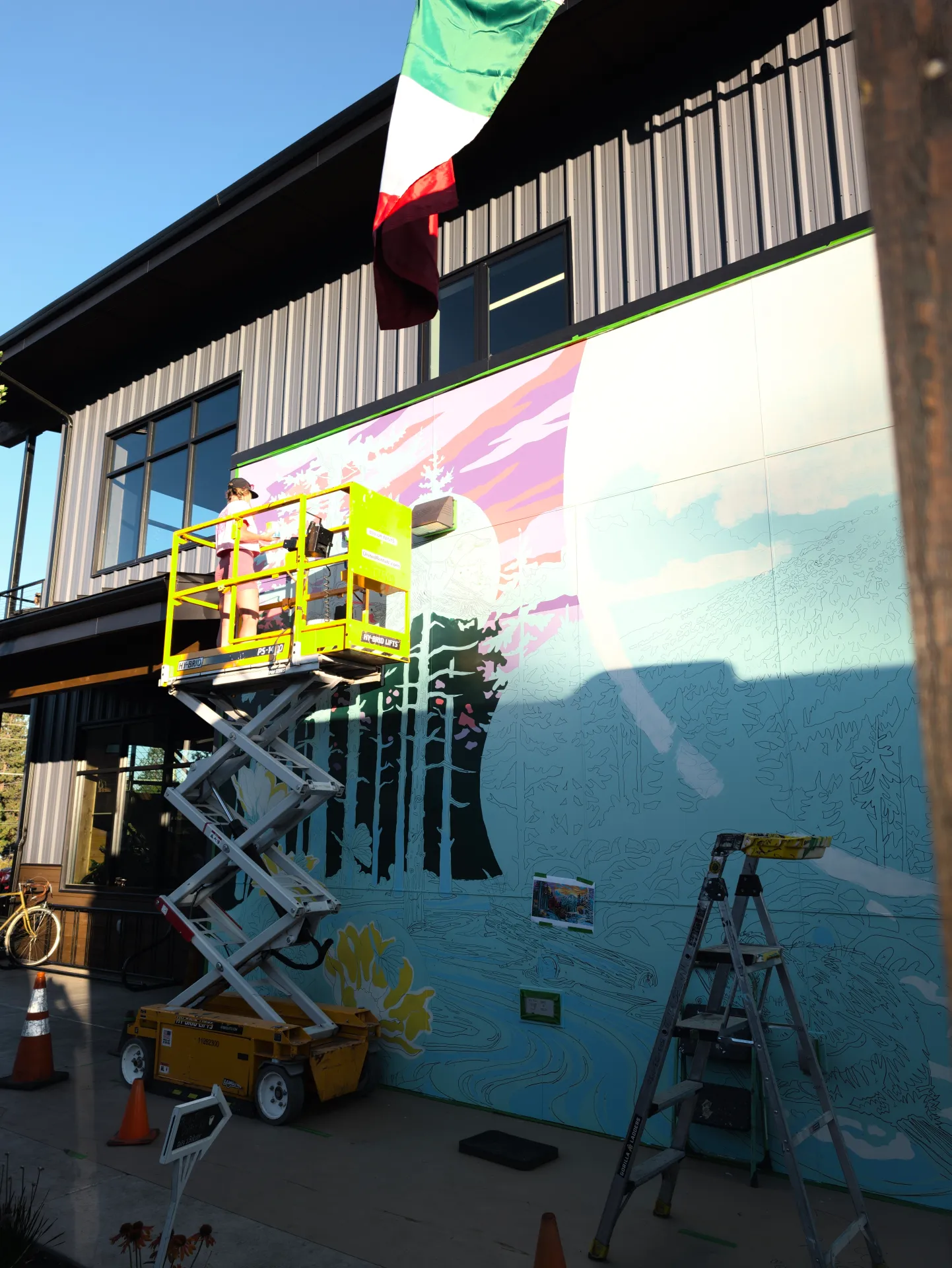 A yellow lift holds an artist painting a vibrant mural of trees and a mountain sunset on an outdoor wall, with a ladder, traffic cones, and bicycle positioned nearby.