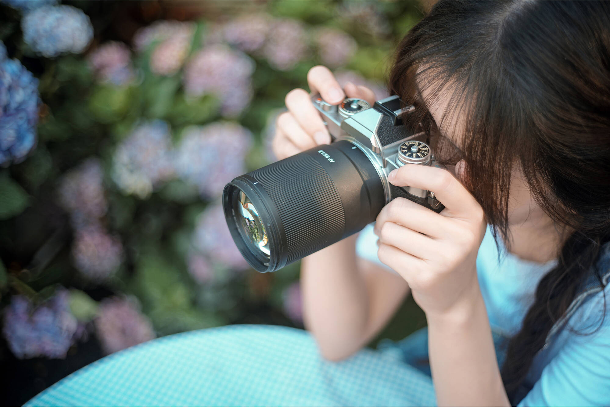 Person in a light blue shirt holding a camera up to their face, with blurry flowers and greenery in the background.