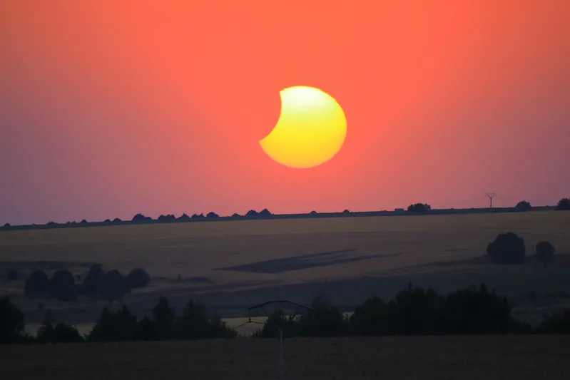 A rolling hill under a bright orange sky. The sun is visible, but with a bite taken out of it by the moon.