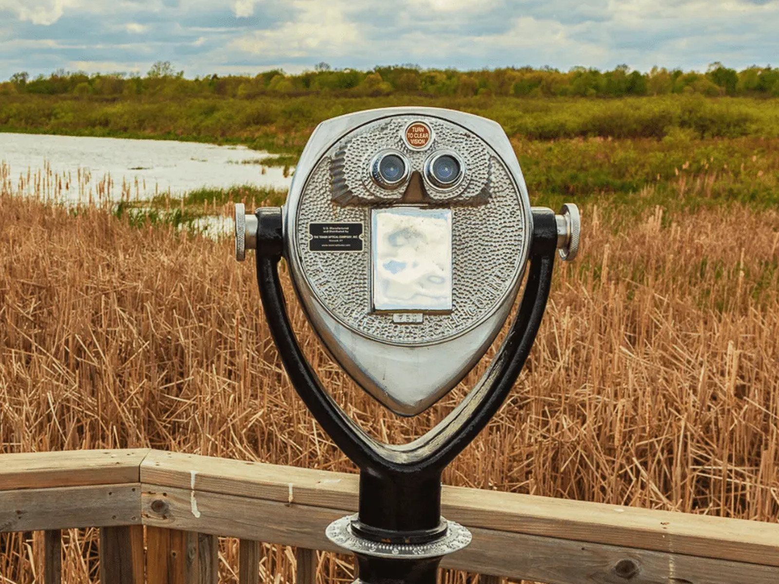 Coin-operated binocular viewer with a metallic finish mounted on a wooden deck, facing a marsh with tall brown grass, water, and green trees beneath a cloudy sky.