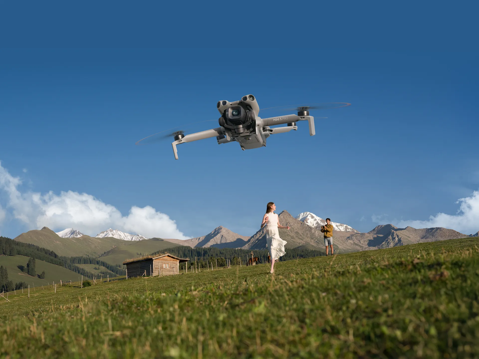 the dji mini 5-pro hovers in a blue sky over a green field in mounains a woman in a white dress runs toward it
