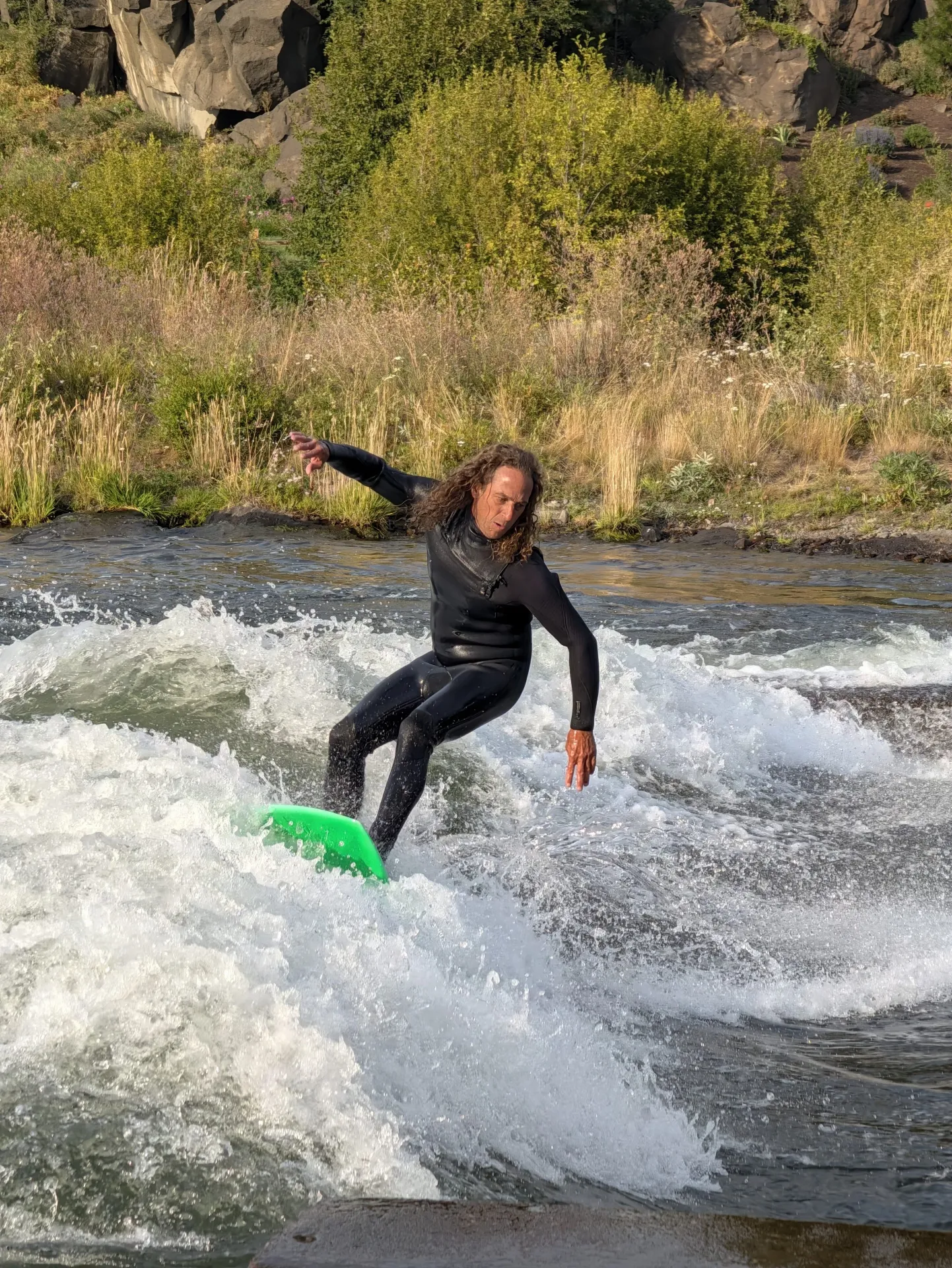 A person in a black wetsuit stands on a bright green surfboard while surfing a river wave, with white water and greenery in the background.