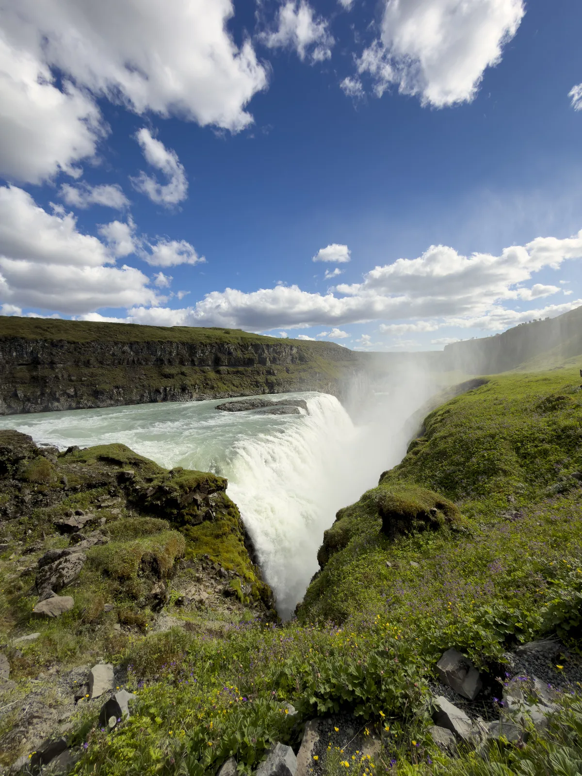 Wide waterfall flowing between green cliffs beneath a bright blue sky with scattered clouds; wildflowers and grass cover the rocky foreground.