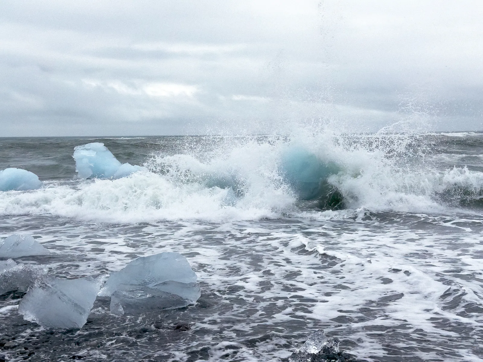 Waves crashing against blue ice chunks floating in a gray, stormy sea, with cloudy skies overhead and additional pieces of ice scattered in the cold water near the shore.