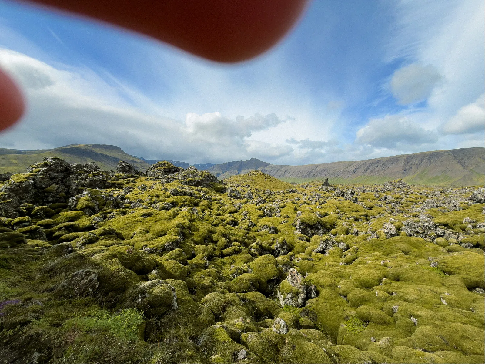 Mossy rocks in a natural landscape with scattered clouds in a blue sky and distant mountains; two fingers partially obscure the top of the view.