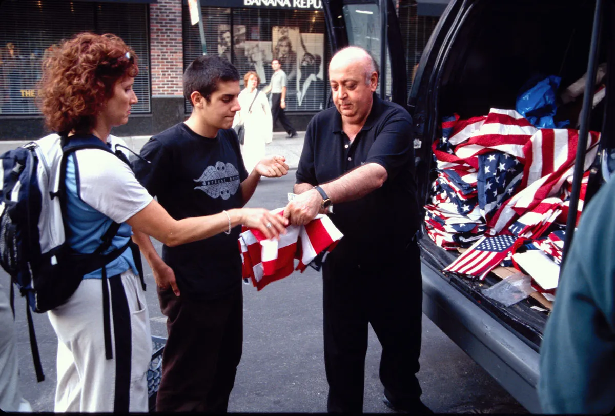 Group stands beside open vehicle trunk filled with folded American flags; a flag is being handed to two others on a city sidewalk.