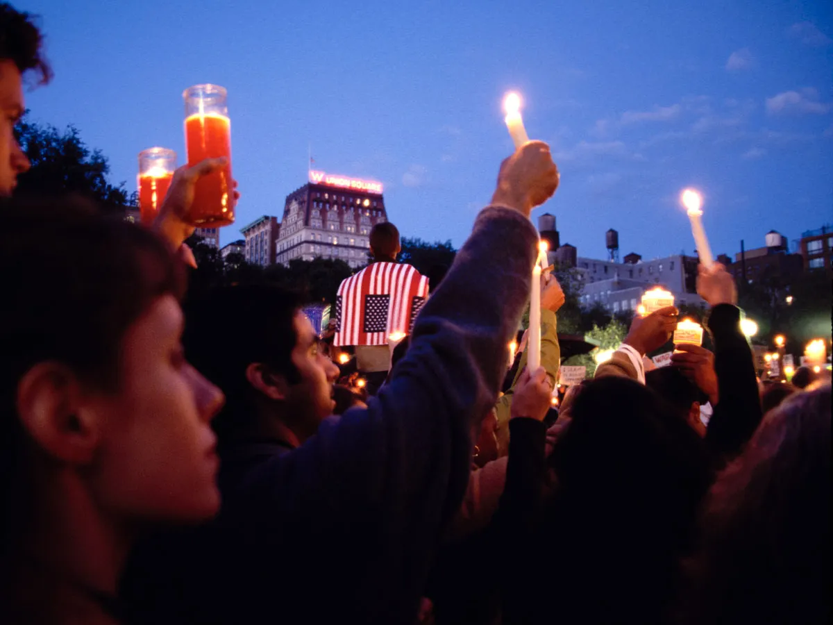 Lit candles held by people at an outdoor evening vigil; one individual wears a jacket with a US flag design. Buildings and a sign reading