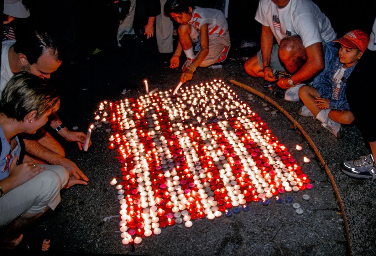 Candles arranged on the ground form an American flag pattern, with a group of people gathered around and lighting the candles during a tribute.