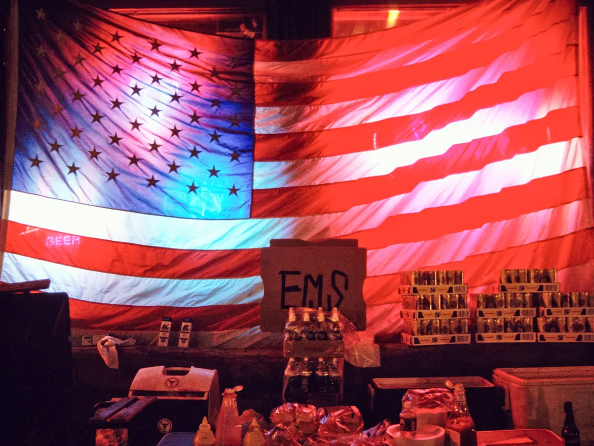 Large American flag serves as a backdrop for a table displaying assorted food items, drinks, condiments, and a cardboard sign marked