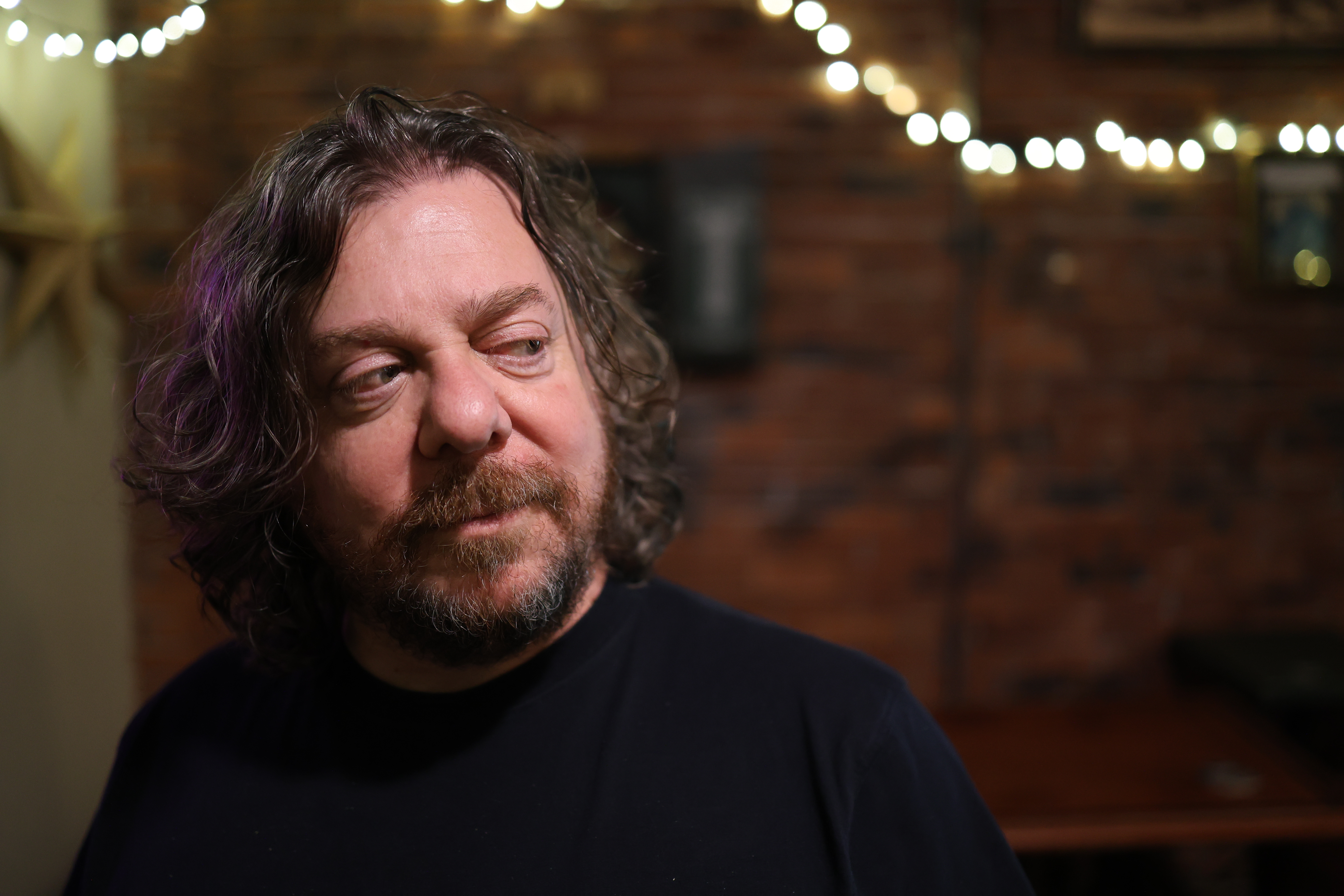 A man with curly hair looks askance at something off to the right of the camera. The scene is in low light with a brick wall and a string of lights in the background