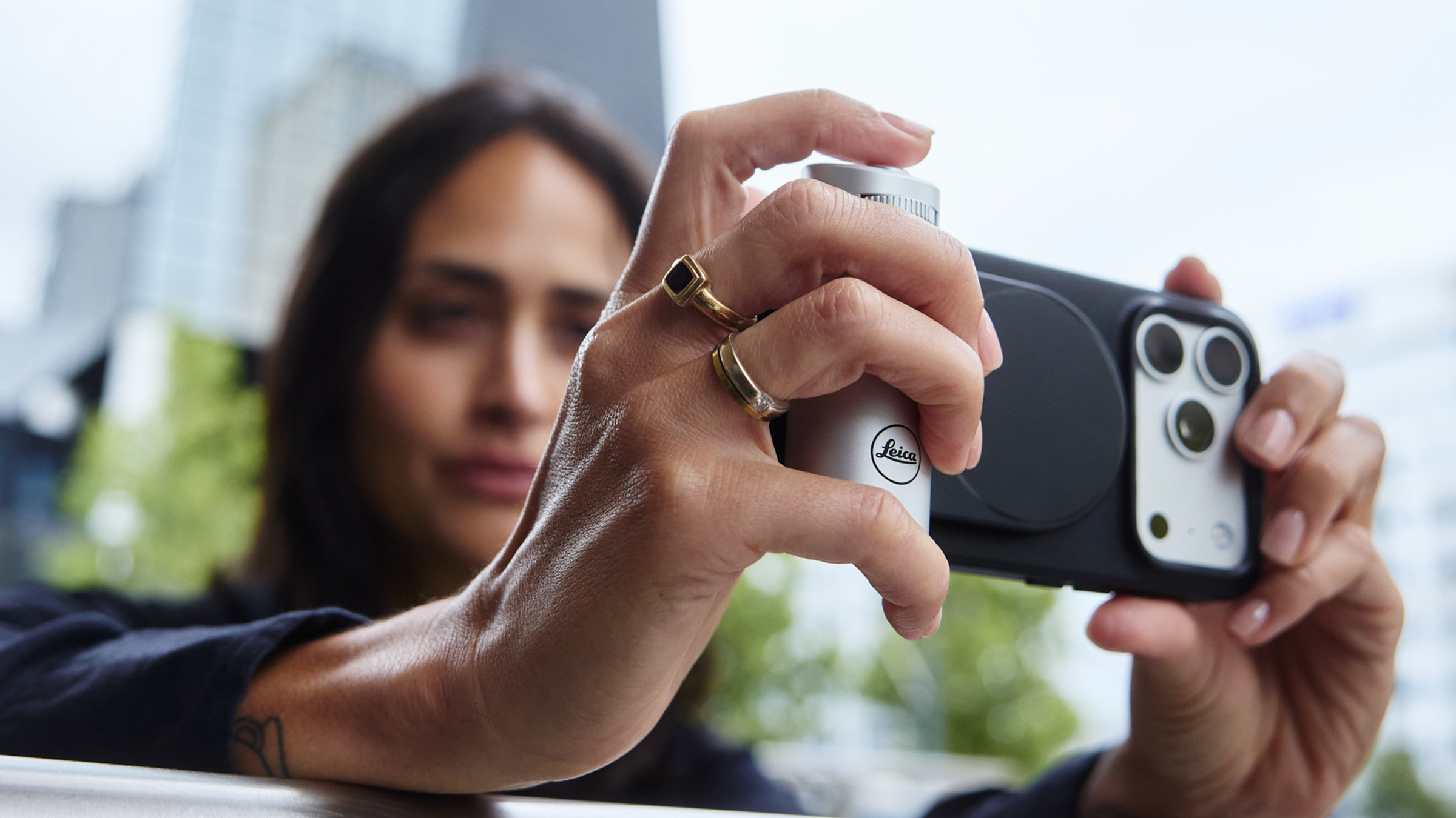 Smartphone with a camera grip attachment held in a hand with rings, aimed outdoors; blurred buildings visible in the background.