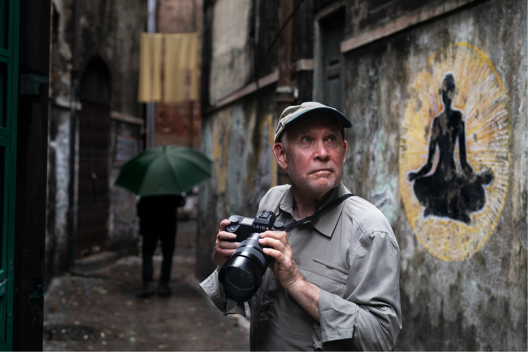 Person holding a camera looks up in a narrow alley with faded wall art; another person is visible in the background with a green umbrella on a rainy day.