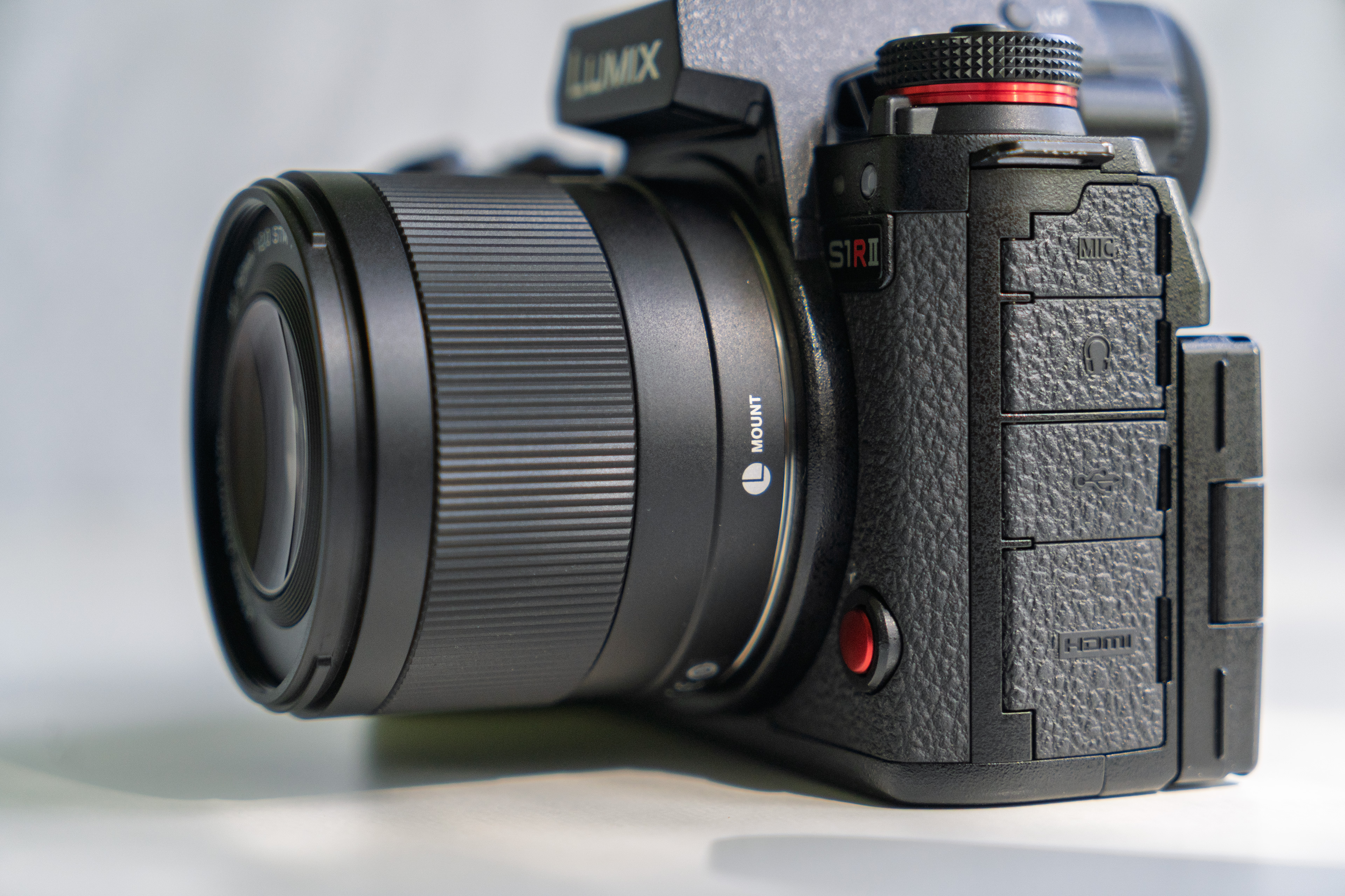 Black Lumix digital camera with visible lens, textured grip, side ports, and a red button, resting on a light surface with a blurred background.