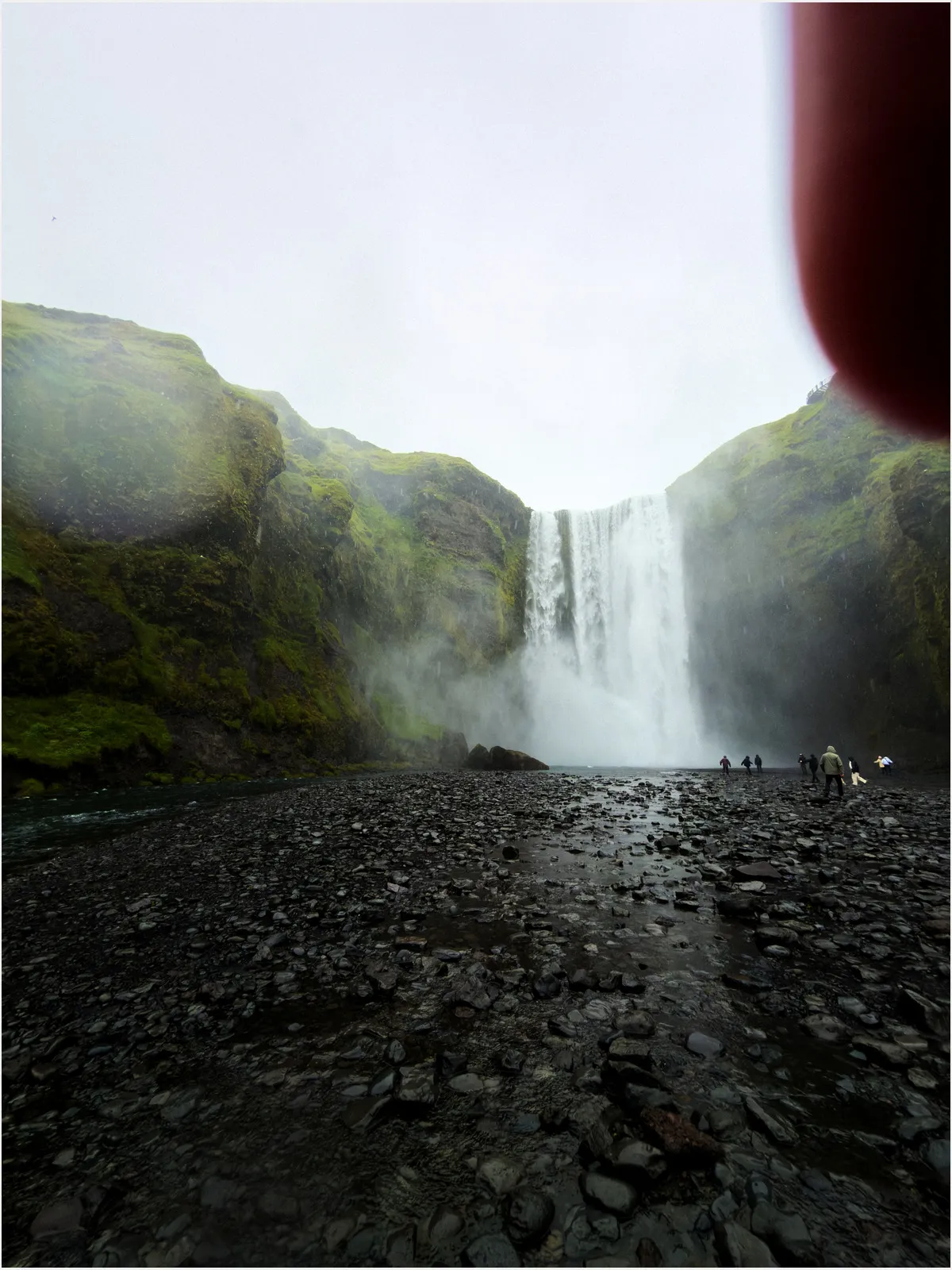 Wide waterfall cascading down mossy cliffs into a rocky riverbed, with several people standing nearby. A red finger partially covers the top right corner. Overcast sky in the background.