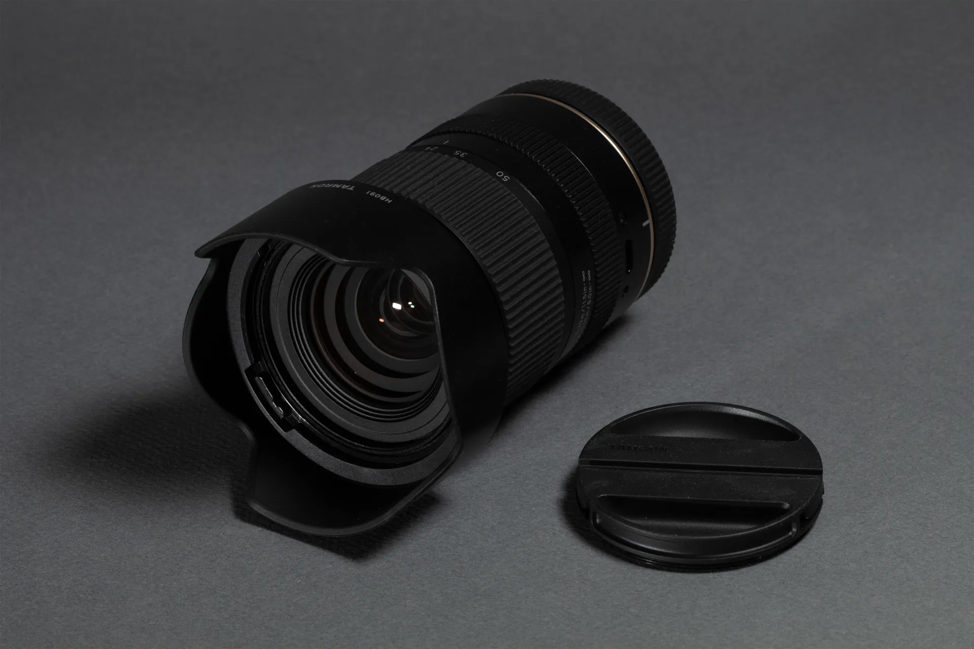 Black Tamron camera lens with hood and detached lens cap on a gray surface.