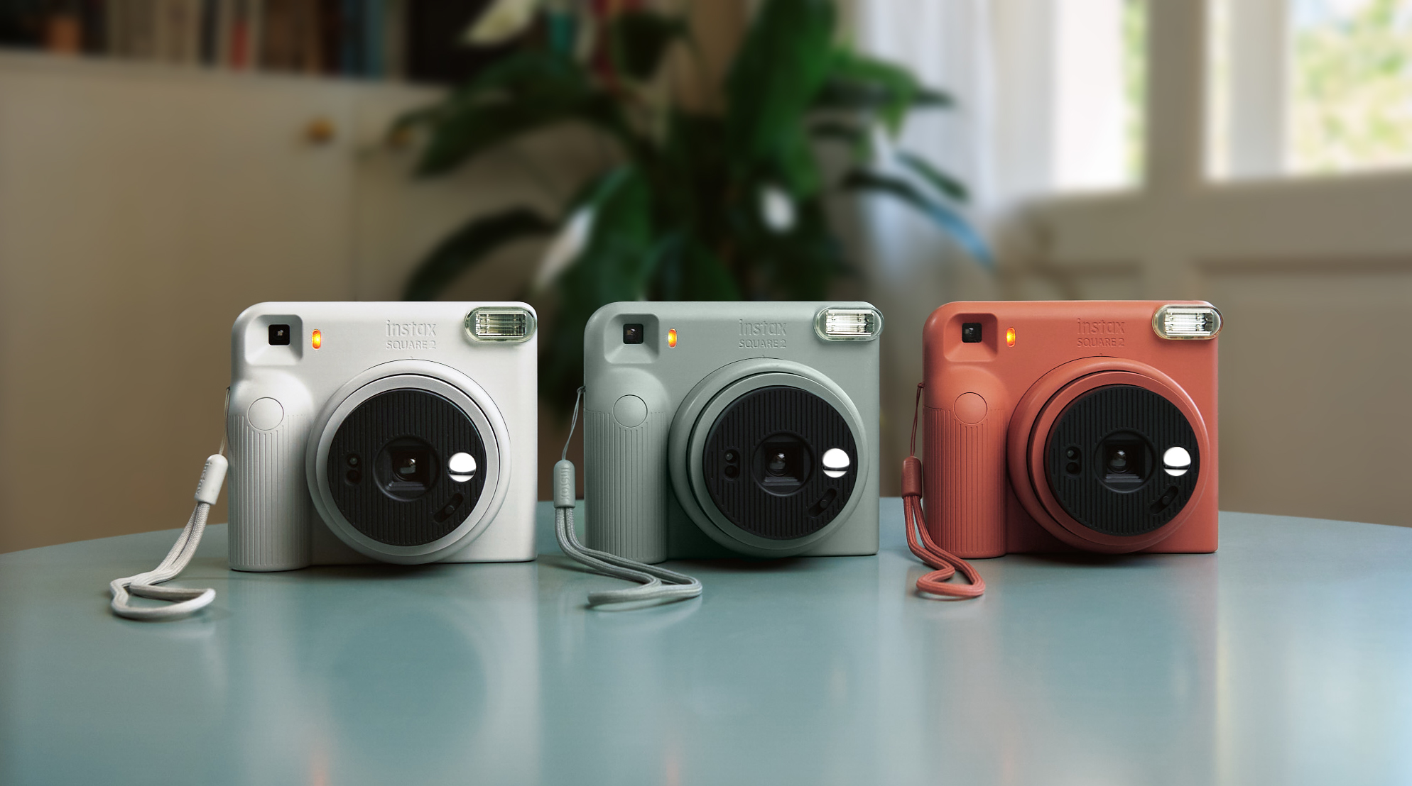 Three Instax Square 2 instant cameras in white, sage green, and terracotta orange, positioned side-by-side on a table.