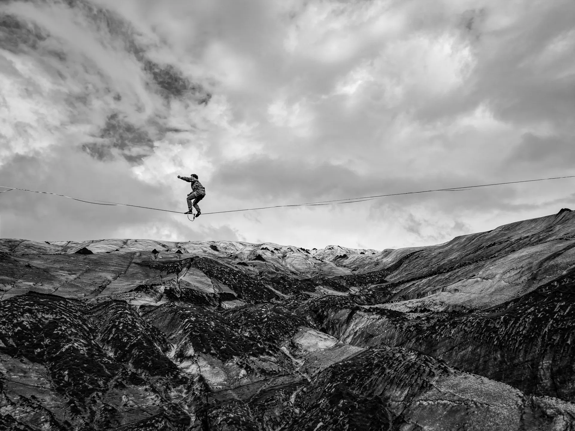 Person balancing on a slackline above a rocky mountain slope under a cloudy sky.