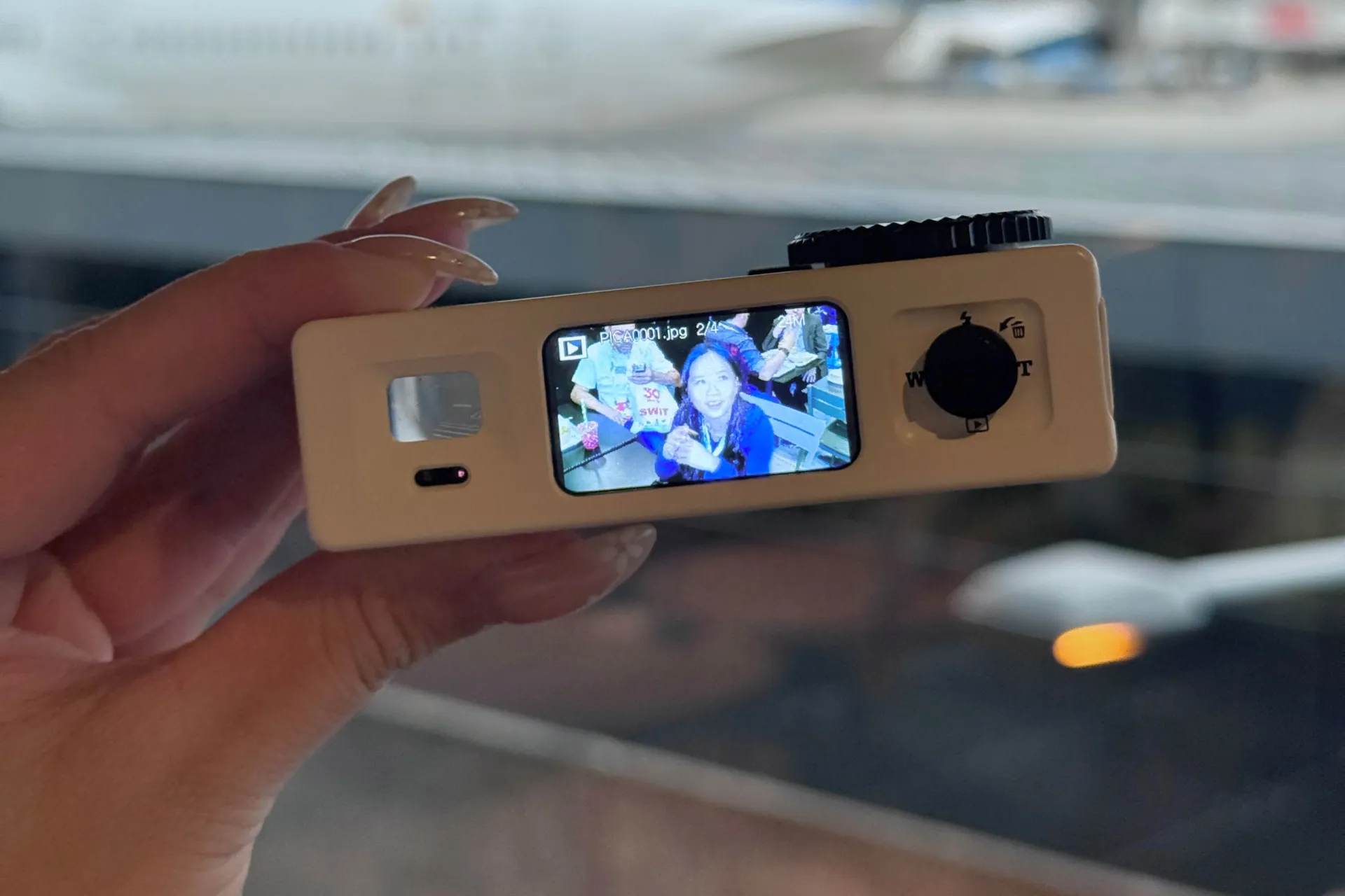 A small digital camera in a hand displays its screen with a selfie of two people, while a blurred airplane and runway appear in the background.