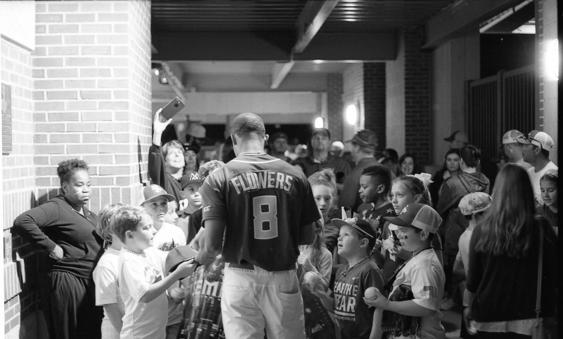 Baseball player in a Flowers jersey signs autographs for a crowd of children in a stadium corridor.